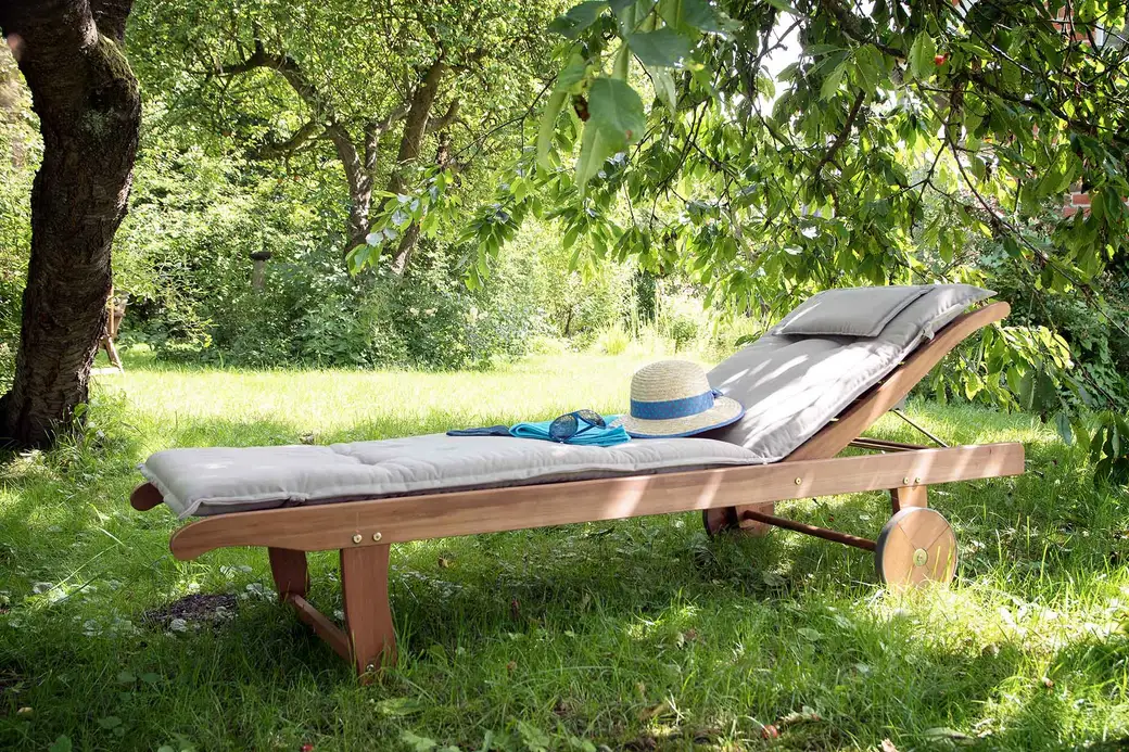 Holz-Gartenliege mit Auflage und Strohhut unter einem Obstbaum im Garten